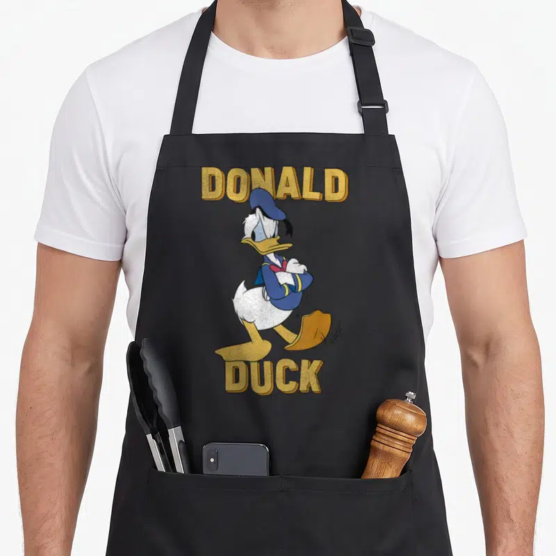Tablier de cuisine noir avec illustration de Donald Duck aux bras croisés et texte Donald Duck en lettres dorées.