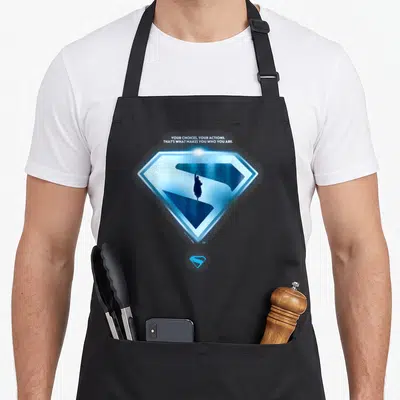 Tablier de cuisine noir avec emblème Superman bleu lumineux et silhouette centrale sous une citation en anglais.