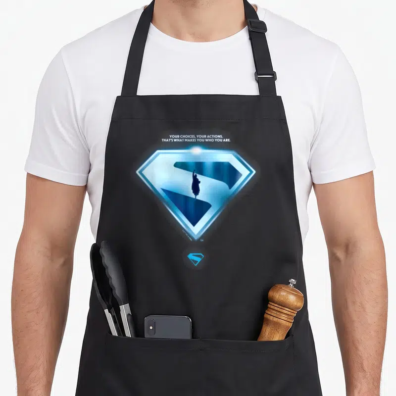Tablier de cuisine noir avec emblème Superman bleu lumineux et silhouette centrale sous une citation en anglais.