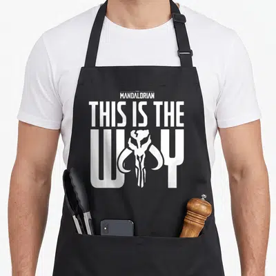 Tablier de cuisine noir avec le logo The Mandalorian, le crâne de mythosaur et le texte This Is The Way en blanc.