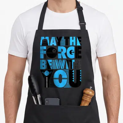 Tablier de cuisine noir avec typographie May The Force Be With You bleue et noire intégrant des icônes Star Wars comme Yoda et Darth Vader.