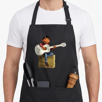 Tablier de cuisine noir avec Miguel Rivera assis sur une caisse jouant d'une guitare blanche ornée de motifs floraux.