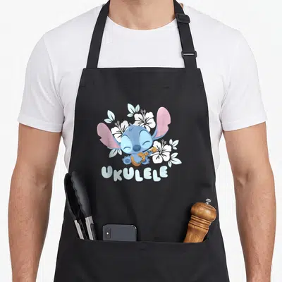 Tablier de cuisine noir avec Baby Stitch jouant de l'ukulele entouré de fleurs blanches et du mot Ukulele imprimé en bas.