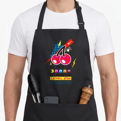 Tablier de cuisine noir avec illustration Ready to Power Up Level 256, Pac-Man et fantômes colorés sur fond géométrique.