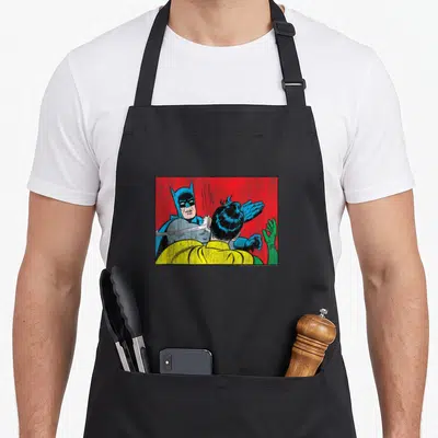 Tablier de cuisine noir avec une illustration colorée de Batman tapant Robin en pleine figure sur fond rouge.