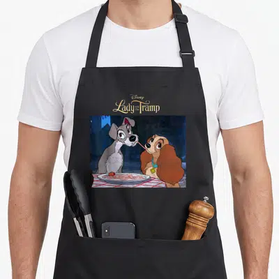 Tablier de cuisine noir avec illustration colorée de Lady et le Tramp partageant un plat de spaghettis sur fond bleu nuit.