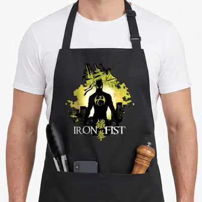 Tablier de cuisine noir avec silhouette Iron Fist, paysage urbain jaune et inscription IRON FIST en blanc.