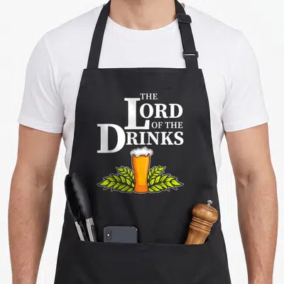 Tablier de cuisine noir avec le texte The Lord of the Drinks au-dessus d'un verre de bière orné de feuilles vertes.