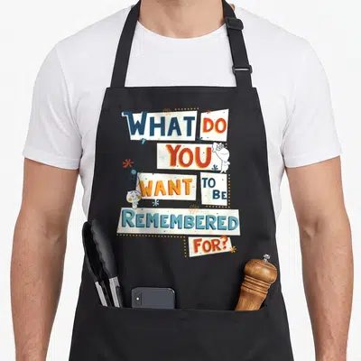 Tablier de cuisine noir avec le texte What Do You Want To Be Remembered For et deux petites entités blanches.