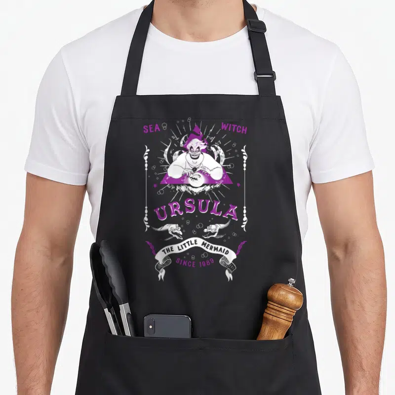 Tablier de cuisine noir avec illustration violette d'Ursula tenant un orbe sous un triangle et le texte Sea Witch.