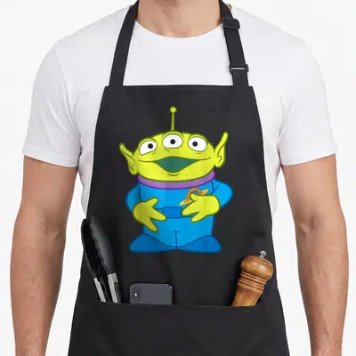 Tablier de cuisine noir avec un alien vert à trois yeux et une combinaison bleue tenant un logo planétaire.