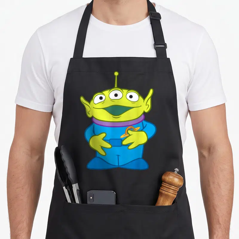 Tablier de cuisine noir avec un alien vert à trois yeux et une combinaison bleue tenant un logo planétaire.