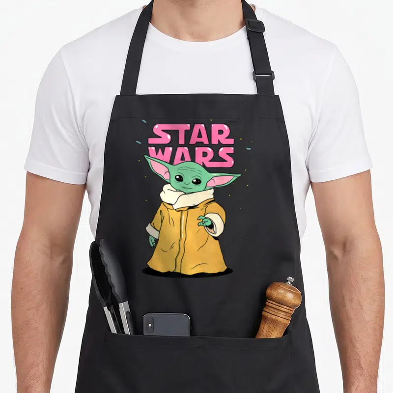 Tablier de cuisine noir avec illustration colorée de Grogu et texte Star Wars en lettres roses bulles.
