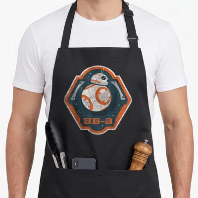 Tablier de cuisine noir avec un badge hexagonal orange et bleu représentant le droïde BB-8 en style vieilli.