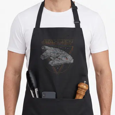 Tablier de cuisine noir avec illustration vintage du vaisseau spatial et logo stylisé en dégradé orange sur fond géométrique.