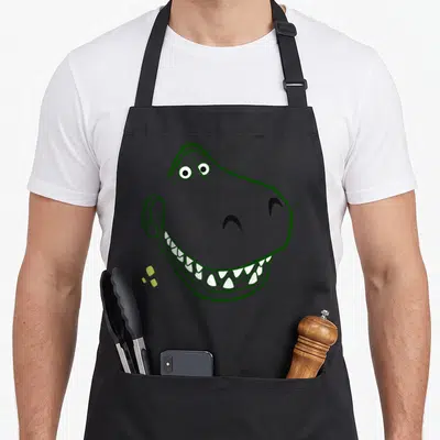 Tablier de cuisine noir avec illustration de Rex dinosaure vert aux dents blanches et yeux ronds.
