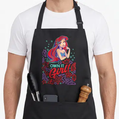 Tablier de cuisine noir avec Ariel aux cheveux rouges et la phrase Own It Girl entourée d'éléments marins et pop.
