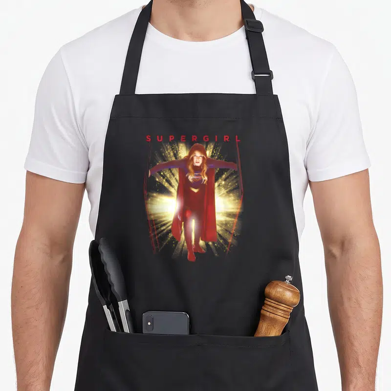 Tablier de cuisine noir avec illustration de Supergirl en plein vol entourée d'éclats lumineux dorés et du titre rouge.