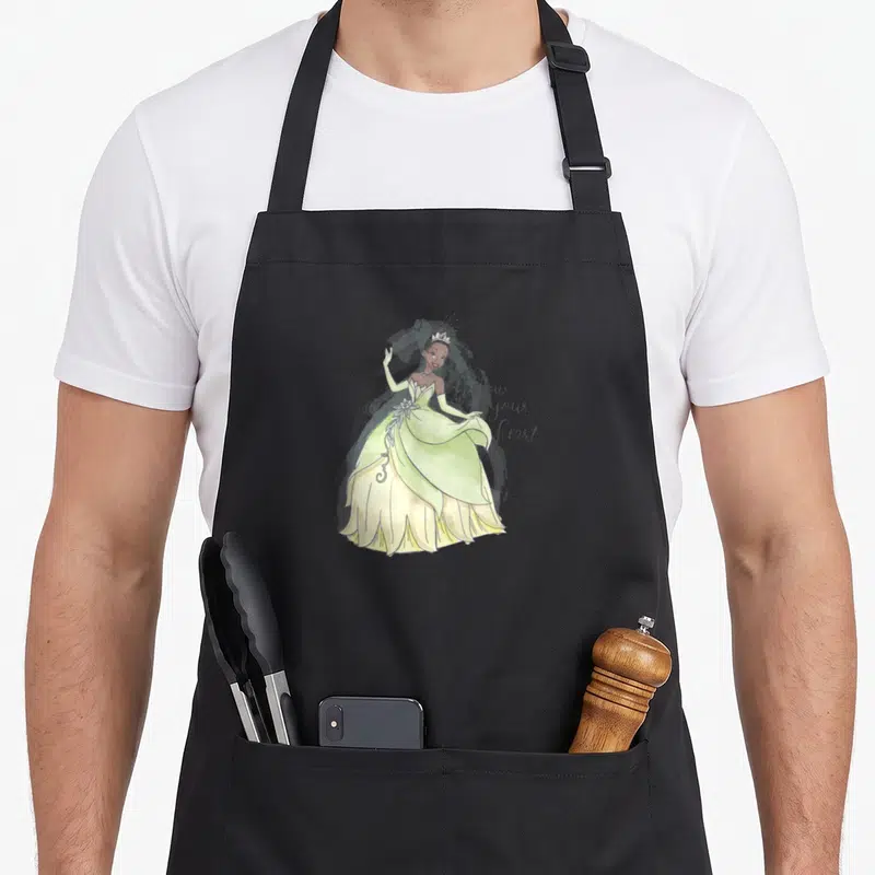 Tablier de cuisine noir avec illustration de Tiana en robe vert clair et texte follow your heart à côté.