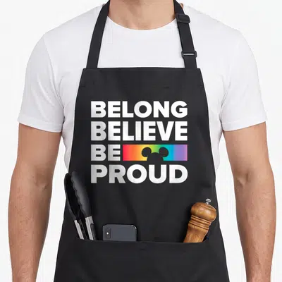 Tablier de cuisine noir avec inscription Belong Believe Be Proud et silhouette arc-en-ciel blanche.