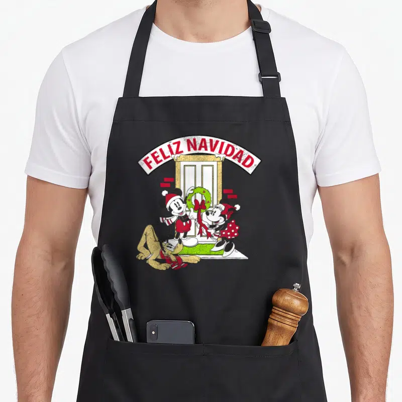 Tablier de cuisine noir avec Mickey, Minnie et Pluto portant des bonnets de Noël devant une porte avec un rameau.