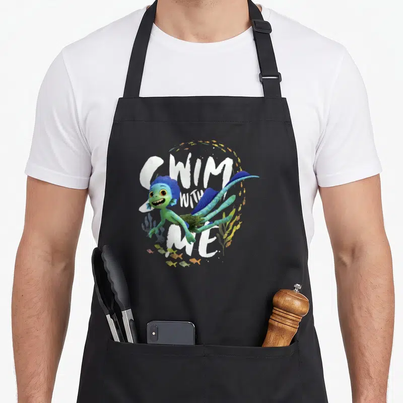 Tablier de cuisine noir avec illustration d'un monstre marin vert nageant et texte blanc Swim With Me entouré de petits poissons.