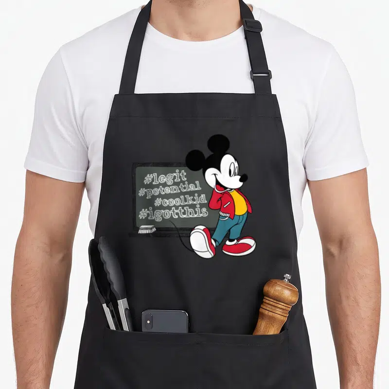 Tablier de cuisine noir avec Mickey Mouse devant un tableau portant des hashtags comme legit et coolkid.