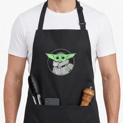 Tablier de cuisine noir avec illustration circulaire de Grogu aux oreilles pointues vertes et col montant gris.