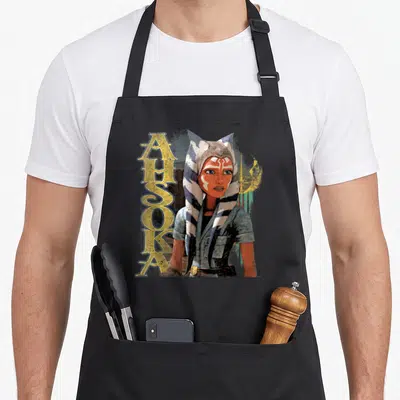 Tablier de cuisine noir avec illustration d'Ahsoka Tano et le texte vertical Ahsoka en lettres dorées stylisées.