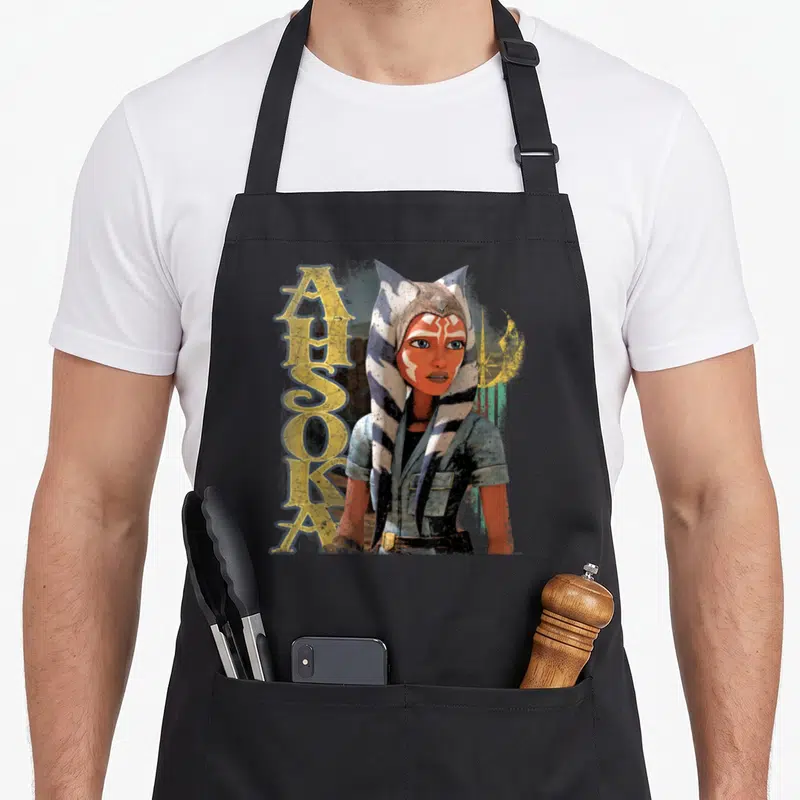 Tablier de cuisine noir avec illustration d'Ahsoka Tano et le texte vertical Ahsoka en lettres dorées stylisées.