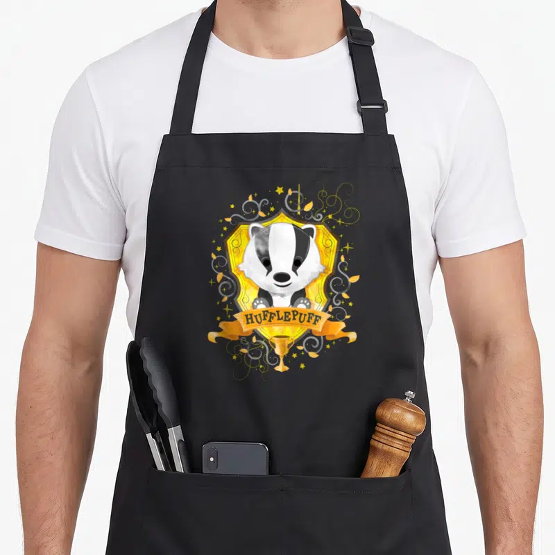 Tablier de cuisine noir avec un blason Hufflepuff montrant un blaireau aquarellé sur fond doré et une bannière orange.