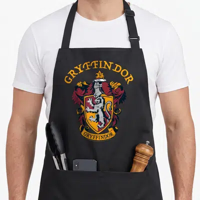 Tablier de cuisine noir avec l'emblème coloré de Gryffindor, un lion blanc sur fond rouge et or, encadré par des plumes et une bannière.