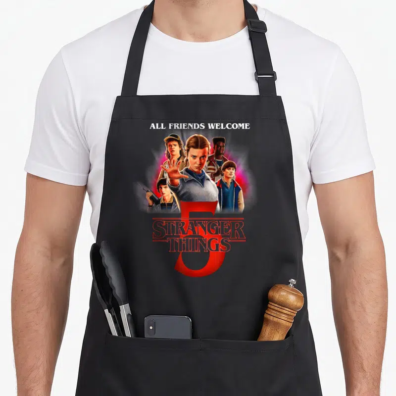 Tablier de cuisine noir avec illustration des personnages et texte All Friends Welcome Stranger Things 5 en rouge.