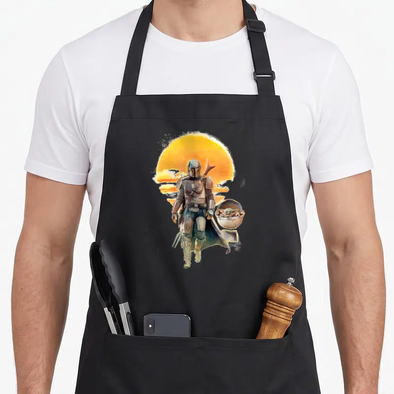 Tablier de cuisine noir avec illustration du Chevalier et The Child devant un soleil couchant orange.