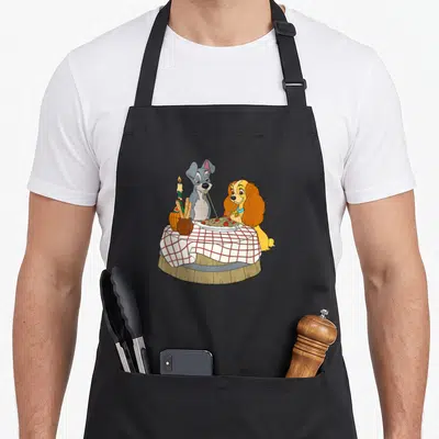 Tablier de cuisine noir avec Lady et Tramp partageant des spaghettis à table.