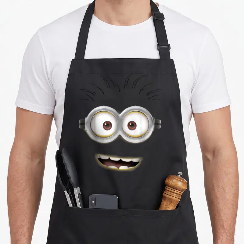 Tablier de cuisine noir avec le visage de Tom Big Face, lunettes et sourire large imprimé au centre.