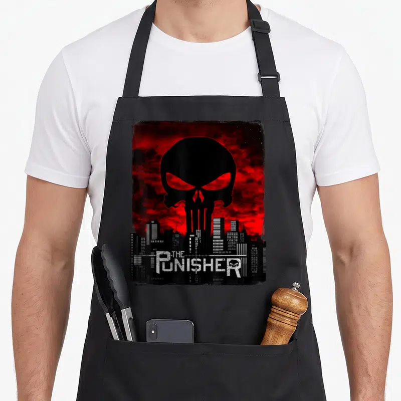 Tablier de cuisine noir avec grand crâne rouge sur fond de ville et logo The Punisher en bas.
