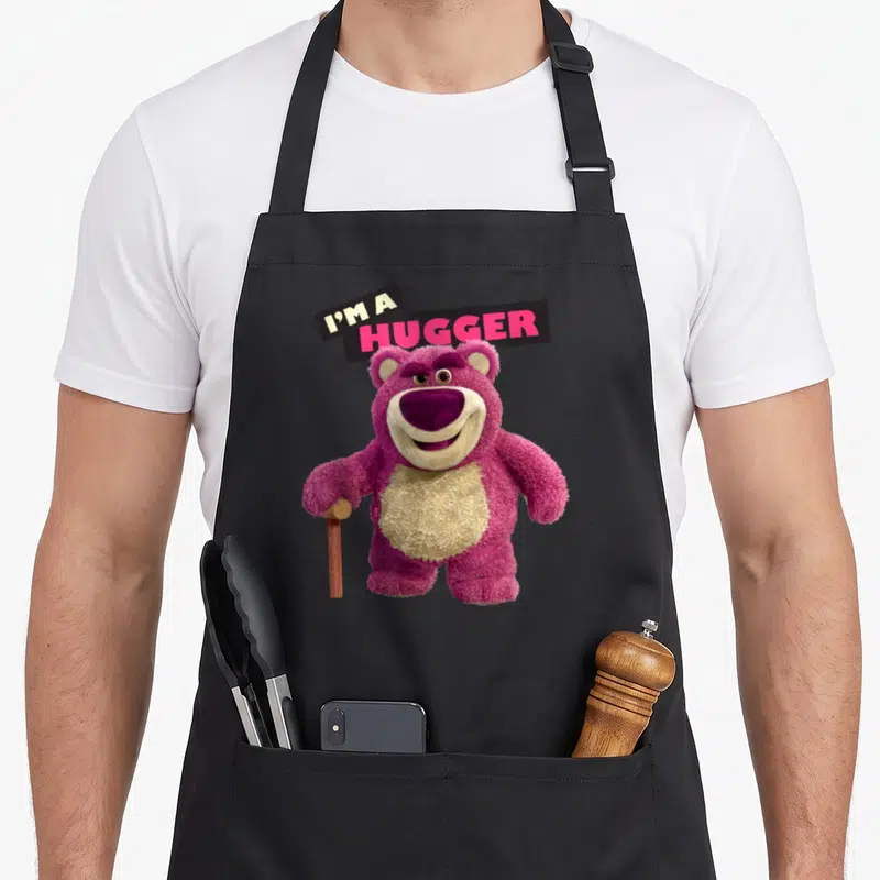 Tablier de cuisine noir avec illustration de l'ours rose Lotso tenant une canne et le texte I'm a Hugger en haut.