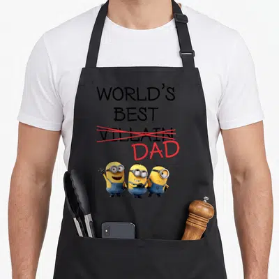 Tablier de cuisine noir avec l'inscription World's Best Villain Dad barrée en rouge et trois Minions souriants en bas.
