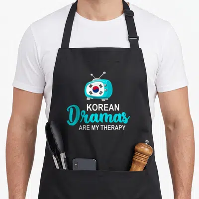 Tablier de cuisine noir avec un téléviseur turquoise affichant le drapeau sud-coréen et une citation humoristique en blanc et bleu.