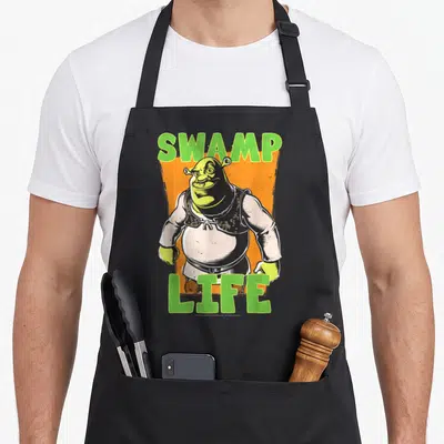 Tablier de cuisine noir avec illustration Shrek cartoon et texte Swamp Life vert sur fond orange texturé.