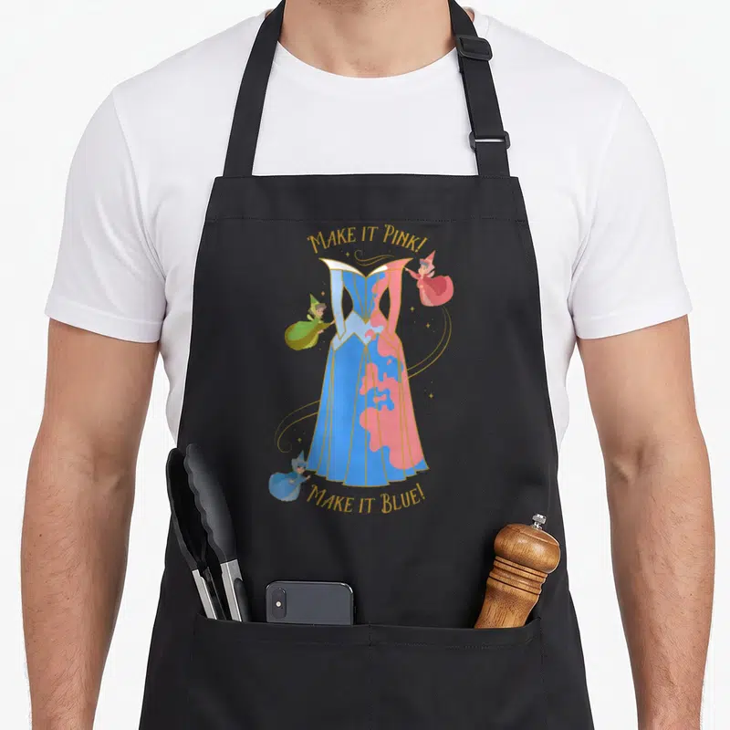 Tablier de cuisine noir avec illustration des trois fées transformant une robe bleue en rose et texte doré Make it Pink Make it Blue.