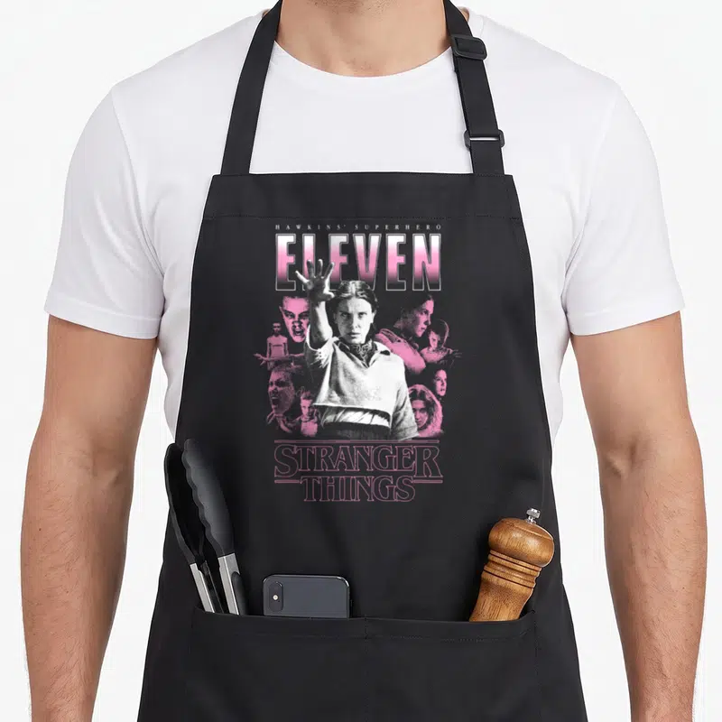 Tablier de cuisine noir avec illustration d'Eleven et texte HAWKINS SUPERHERO ELEVEN STRANGER THINGS en rose et blanc.