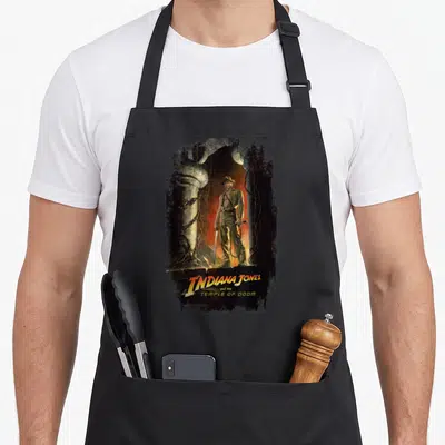 Tablier de cuisine noir avec Indiana Jones debout devant un temple maudit, sabre et whip en main, style poster vintage.
