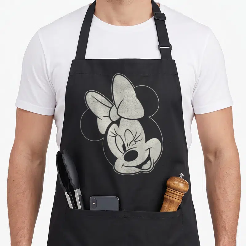 Tablier de cuisine noir avec illustration monochrome du visage de Minnie Wink Face et son grand nœud.
