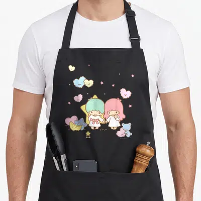 Tablier de cuisine noir avec deux personnages aux cheveux pastel tenant la main sous des étoiles colorées.