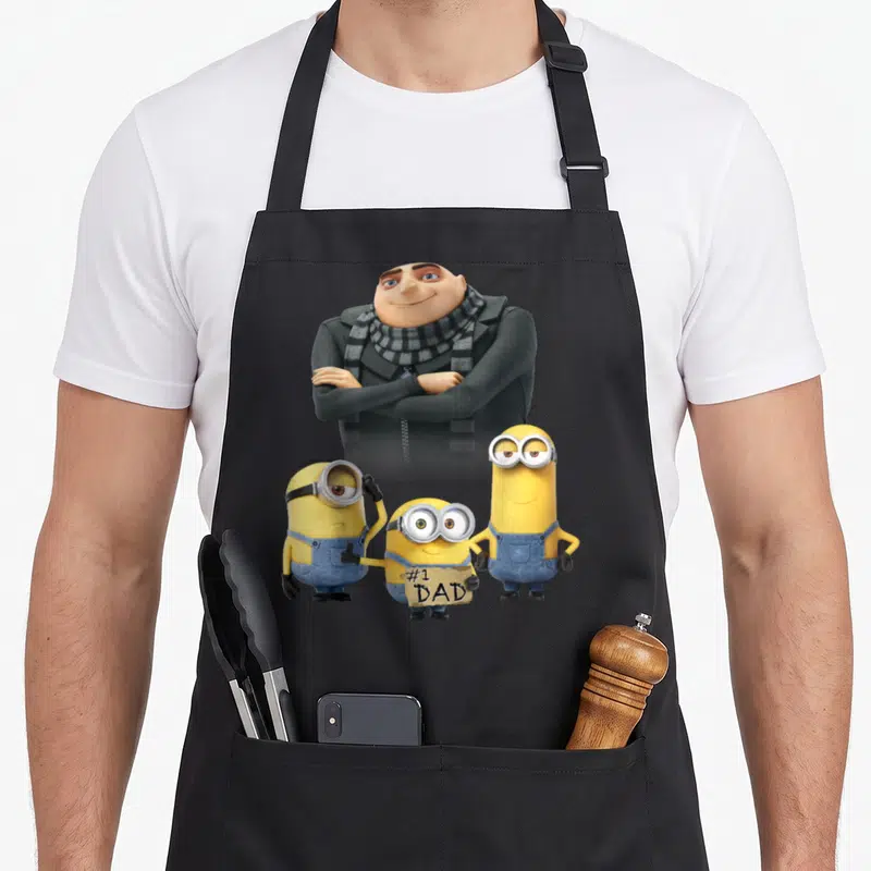 Tablier de cuisine noir avec Gru en fond et trois Minions jaunes, dont un tenant une pancarte #1 DAD.