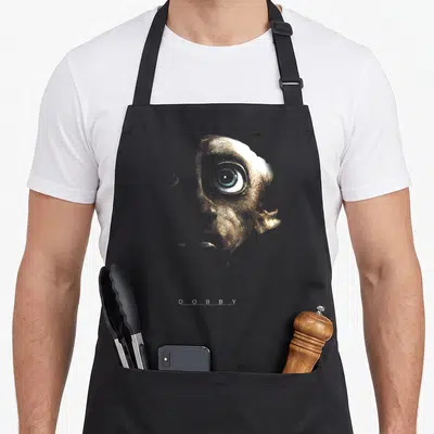 Tablier de cuisine noir avec portrait doré de Dobby, œil bleu distinctif et texte blanc DOBBY en bas du motif imprimé.