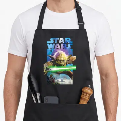 Tablier de cuisine noir avec illustration colorée de Yoda brandissant un sabre laser vert sous le logo Star Wars en lettres bleues spatiales.