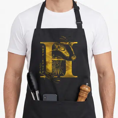 Tablier de cuisine noir avec une grande lettre H dorée et un badger stylisé intégré au design typographique.